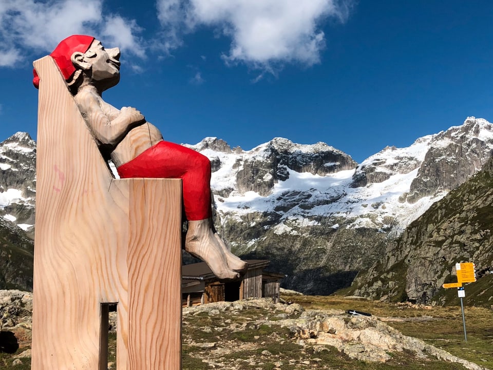 Eine Puppe hockt auf einen überdimensionalen Stuhl, dahinter die Bündner Berge.