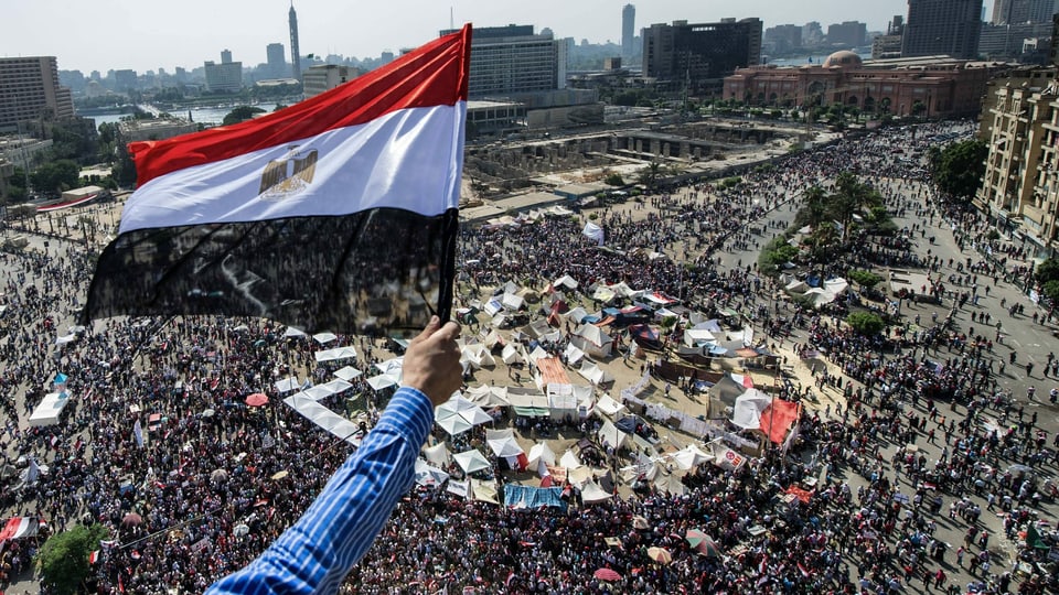Blick auf den Platz, der voll mit Menschen ist. Im Vordergrund ein Arm, der eine ägyptische Flagge hält.