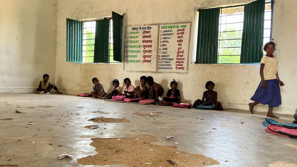 Kinder sitzen auf dem Boden in einer Schule unter zwei grossen Tafeln, auf denen die Wochentage und Monate stehen.