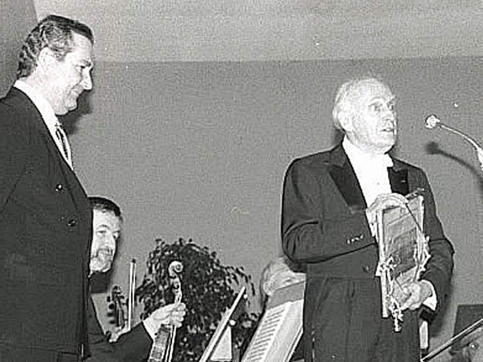 Ein altes Schwarzweissfoto. Links im Bild Boris Banga, rechts spricht Yehudi Menuhin in ein Mikrofon. Im Hintergrund sitzt ein Mann mit einer Geige in der Hand. 
