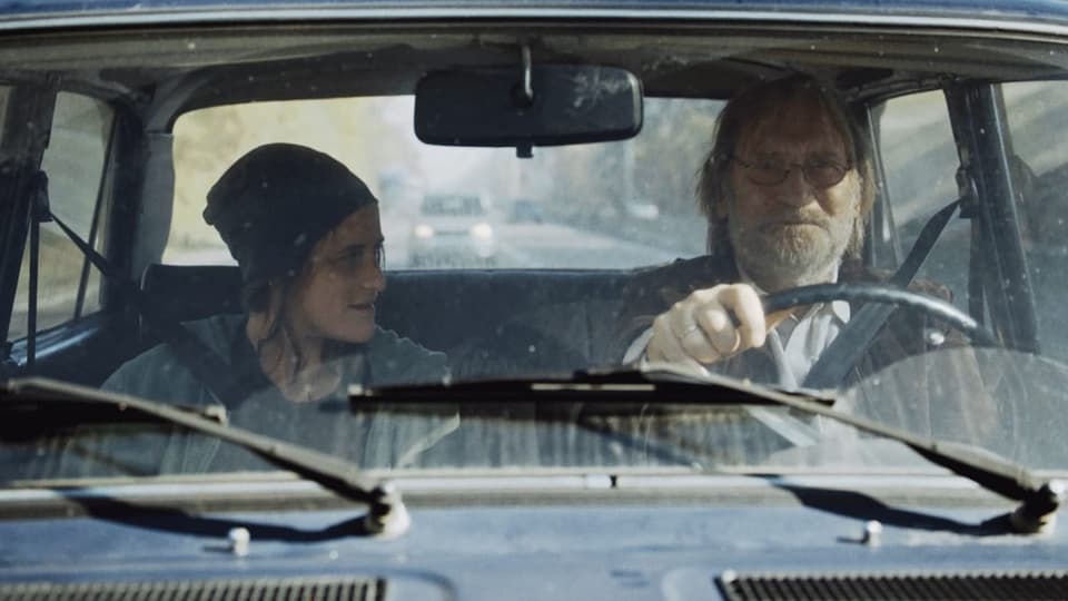 Filmszene_ Eine Frau und ein älterer Mann sitzen in einem Auto.