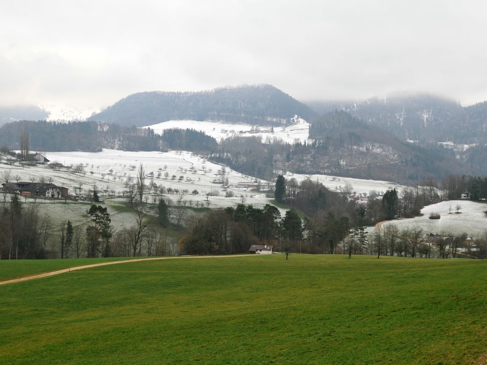 Eine Hügellandschaft mit Schnee in erhöhten Lagen und grünen Wiesen weiter unten.
