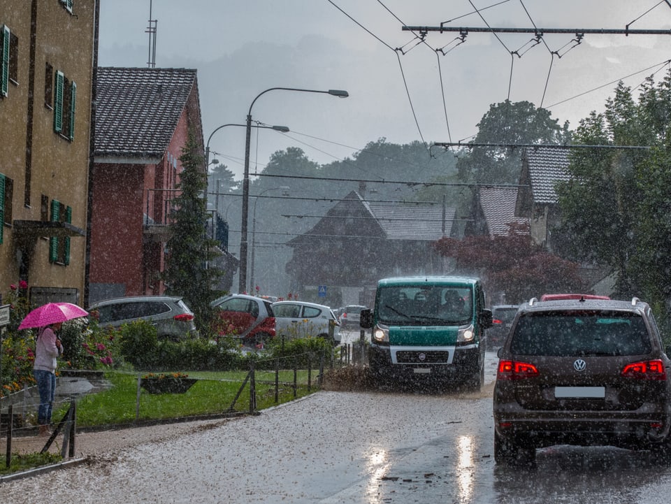 Von braunem Wasser überflutete Strasse. Es regnet weiter, Autos versuchen zu fahren, eine Frau mit einem rosa Regenschirm kämpft sich auf dem Gehweg durch das Unwetter. 