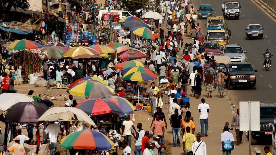 Menschen sind an einem Markt in Guinea-Bissau, es hat viele farbige Sonnenschirme.