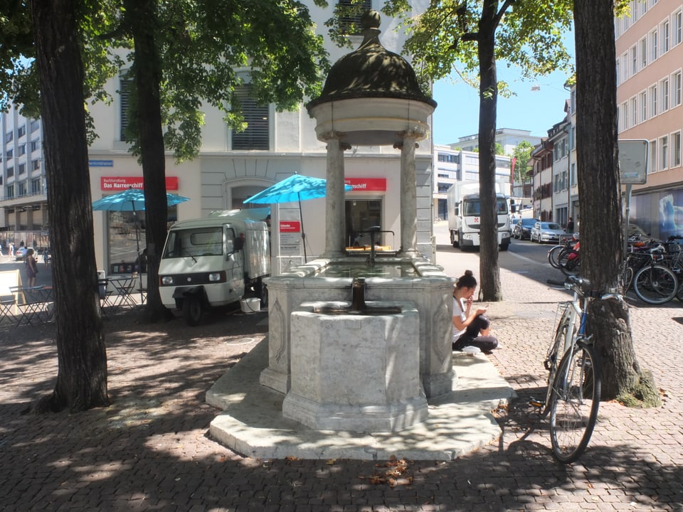 Steinerner Brunnen mit einer kleinen Kuppel über dem Hahnen, neben dem Brunnen stehen vier Lindenbäume. Eine junge Frau sitzt am Brunnenrand und liest. 