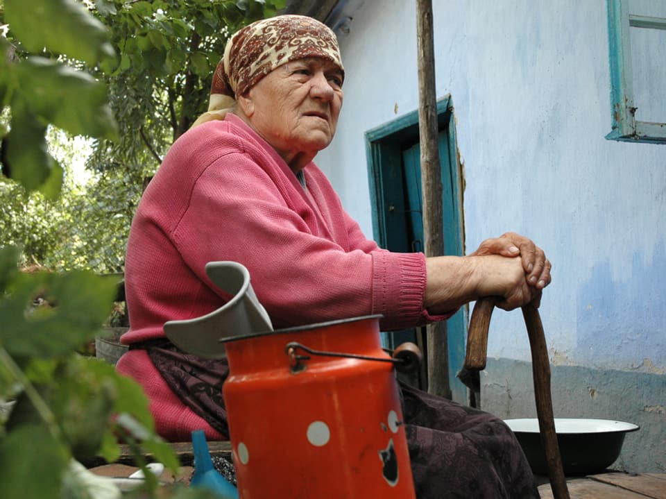 Eine alte Frau sitzt draussen auf einem Stuhl und stützt ihre Hände auf einem Stock ab.