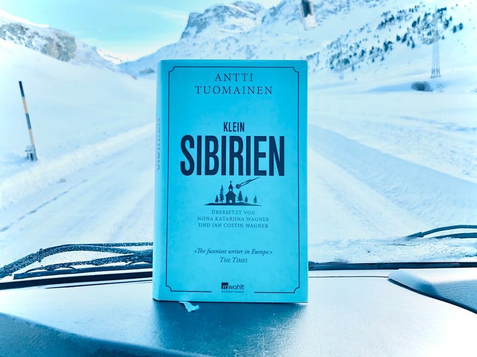 Der Krimi «Klein-Sibirien» von Antti Tuomainen ist vor die Windschutzscheibe eines Autos gehalten. Im Hintergrund sieht mein eine verschneite Strasse.