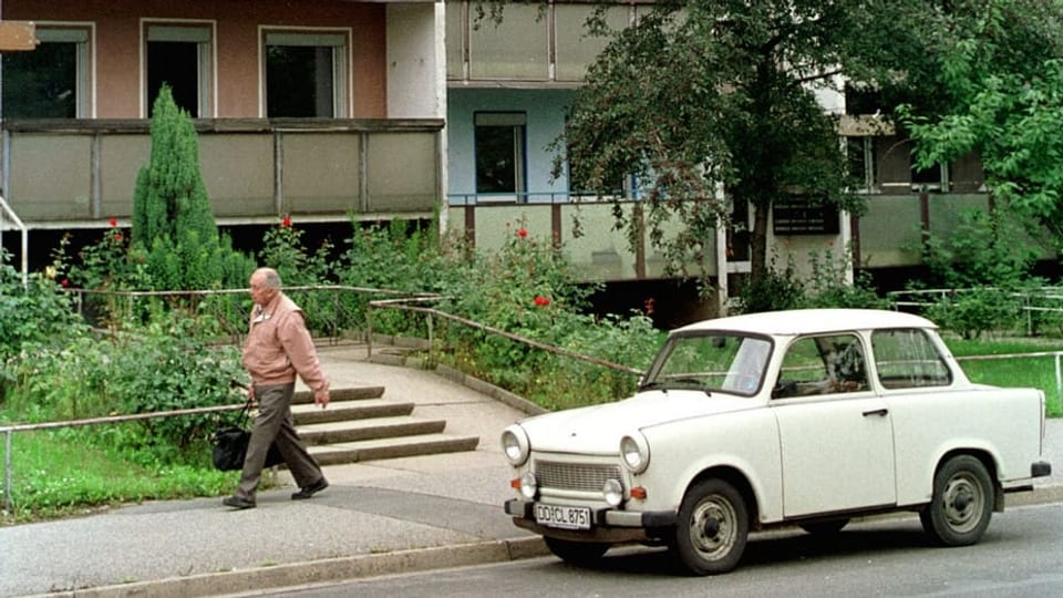 Ein Mann geht vor einem DDR-Plattenbau eine Strasse entlang. Am Strassenrand steht ein Trabant.