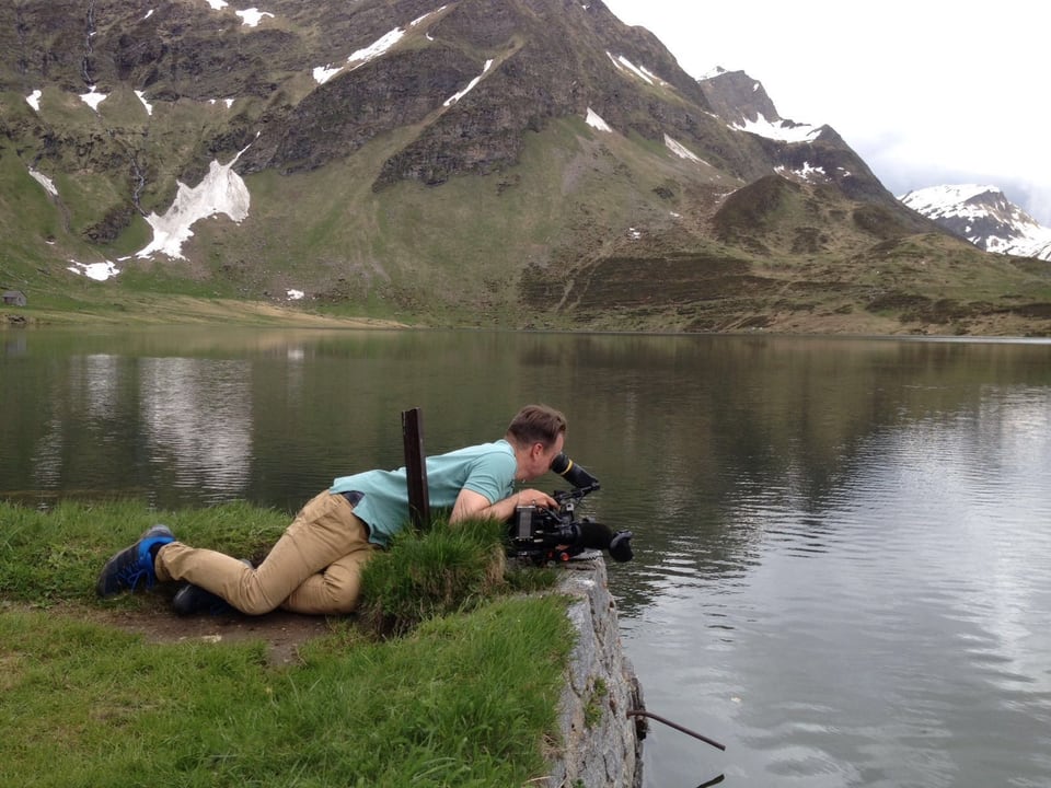 Kameramann liegt am Ufer eines Bergsees am Boden im Gras und filmt. 
