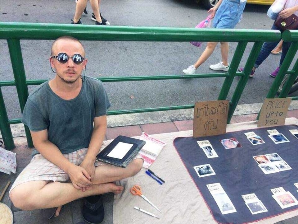 Ein junger Mann mit Sonnenbrille sitzt im Schneidersitz auf dem Boden. Neben ihm liegen Postkarten.