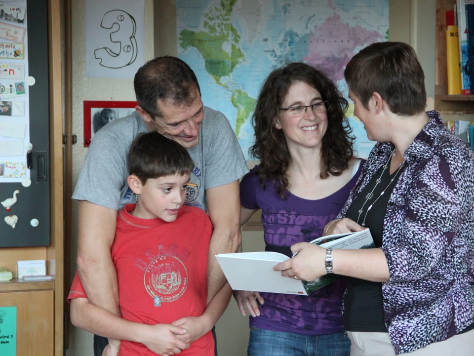 Die Lehrerin zeigt den Eltern von Daniele Ambrosini ein Buch und erklärt etwas.