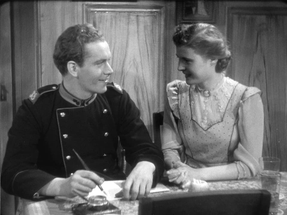 Ein Soldat und eine Frau (Gilberte) sitzen nebeneinander am Tisch und schauen sich an. Der Soldat hält einen Füllfederhalter und hat ein Blatt Papier vor sich.