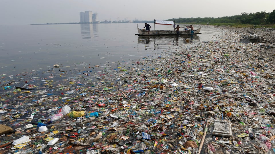 Müll schwimmt am Strand, im Hintergrund ein Boot und noch weiter hinten Hochhäuser.