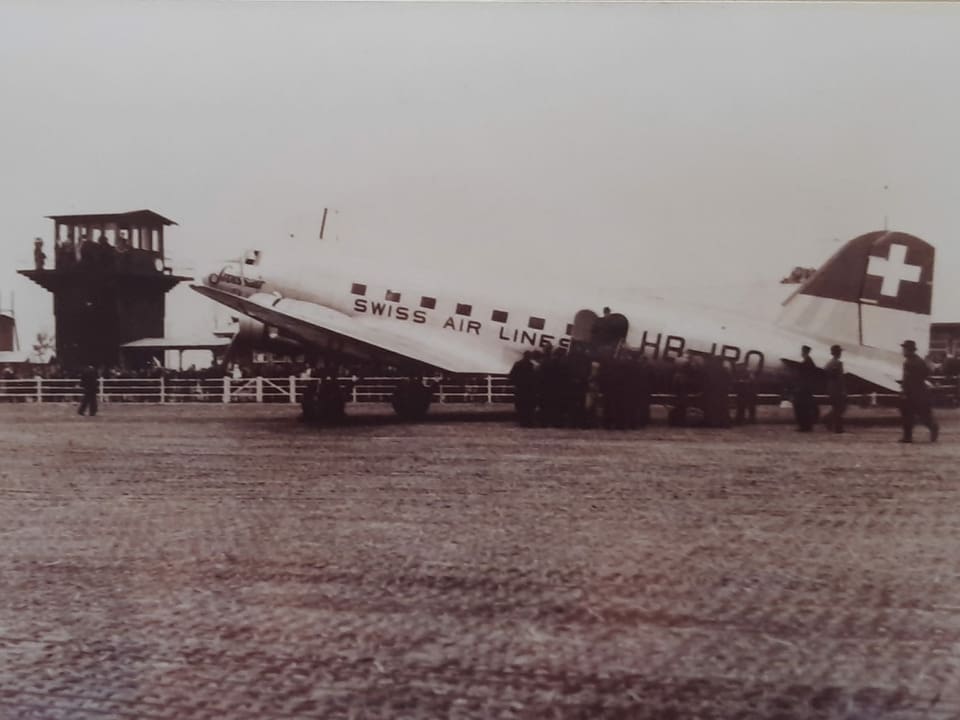 Auf einem historischen Foto steht das Flugzeug DC3 vor einem Kontrollturm.