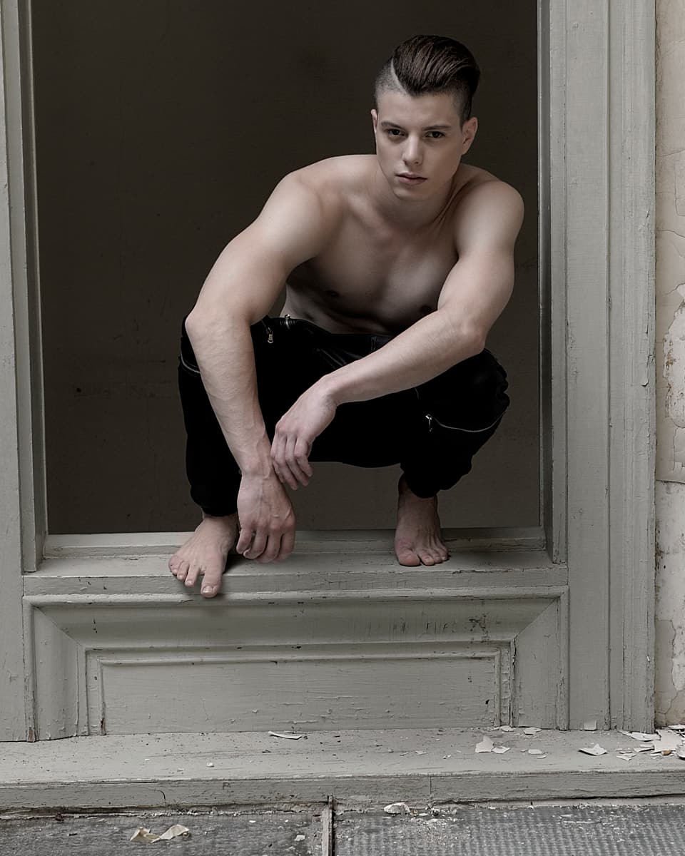 Lucas Fischer posiert kniend in einem Türrahmen. Er trägt eine schwarze Hose und sonst nichts.