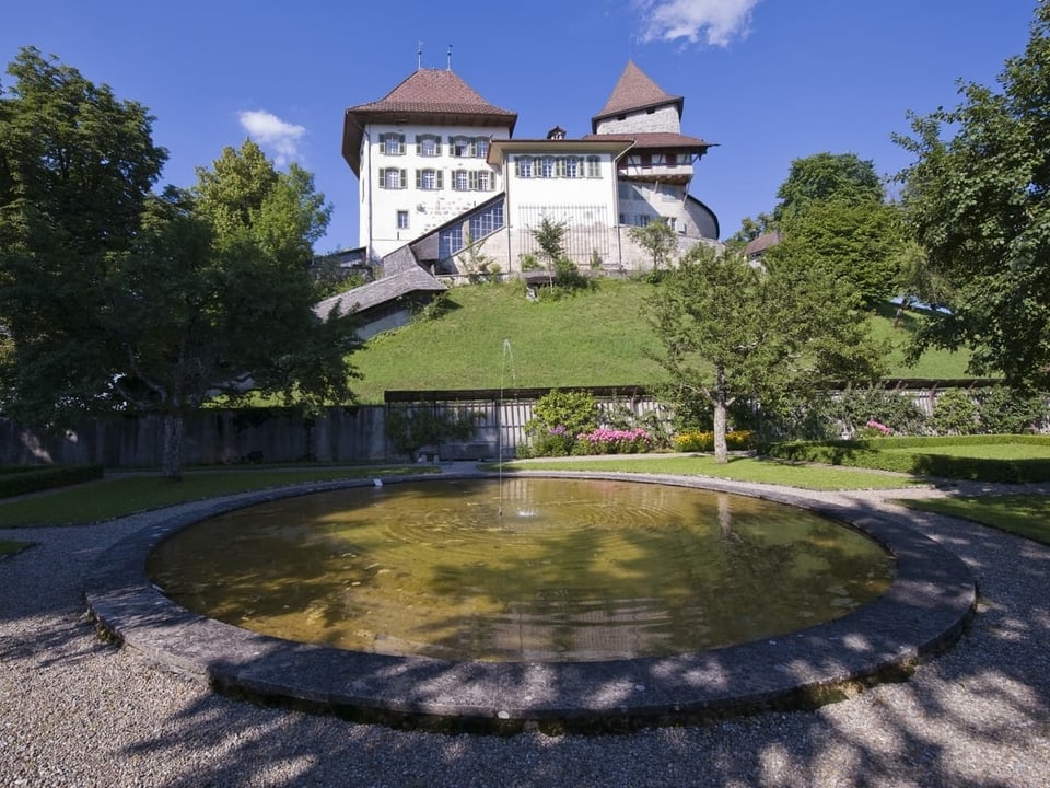 Ein Schloss auf einem grünen Hügel, davor ein künstlicher Brunnen in einer Parkanlage.