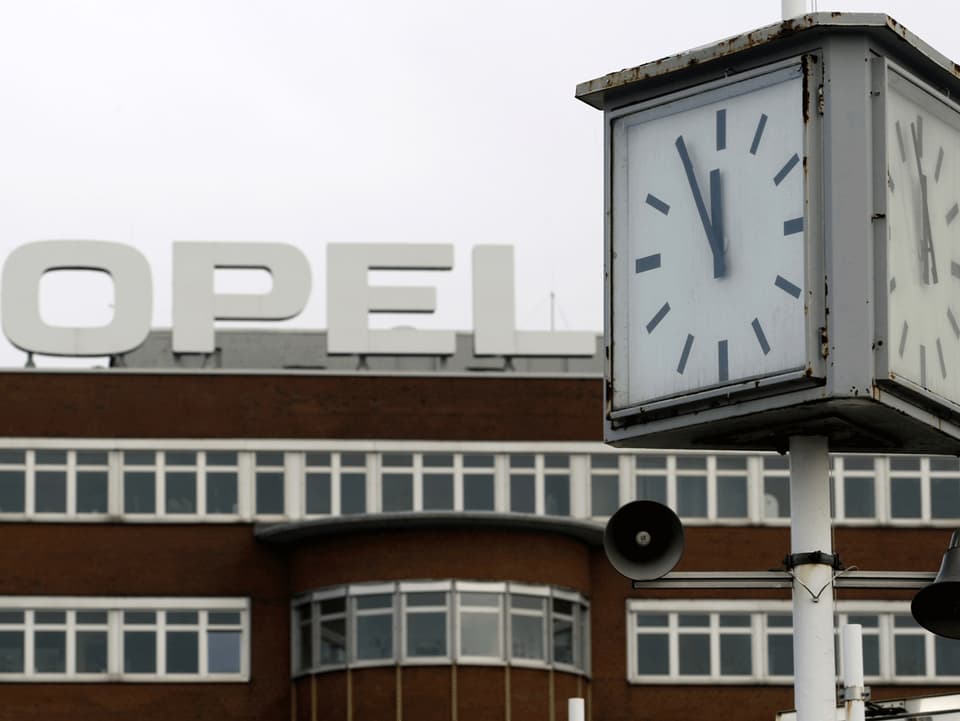 Vor der braunen Fassade des Opel-Werks in Bochum zeigt eine Uhr fünf vor zwölf.