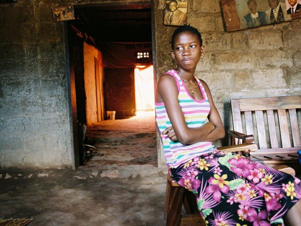 Ein junges schwarzes Mädchen sitzt in einem Steinhaus. Sie hält die Arme vor dem Bauch verschränkt und schaut aus dem linken Bildrand heraus.