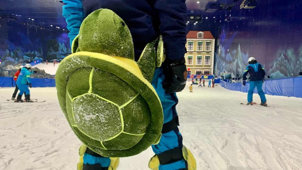 Auf einer Skipiste in der Halle fahren Menschen. Eine Kulisse aus europäischen Häusern ist im Hintergrund zu sehen.  