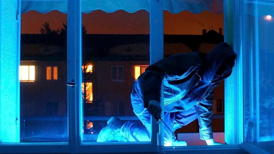 Ein Einbrecher mit Kapuze steigt bei Nacht durch ein Fenster in eine Wohnung.