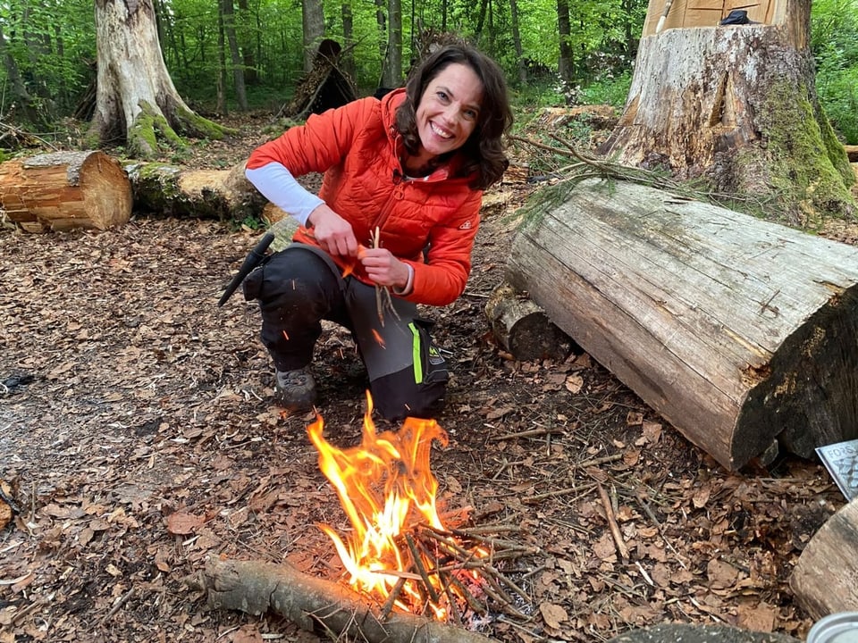 Frau mit Jacke macht auf dem Waldboden ein Feuer.