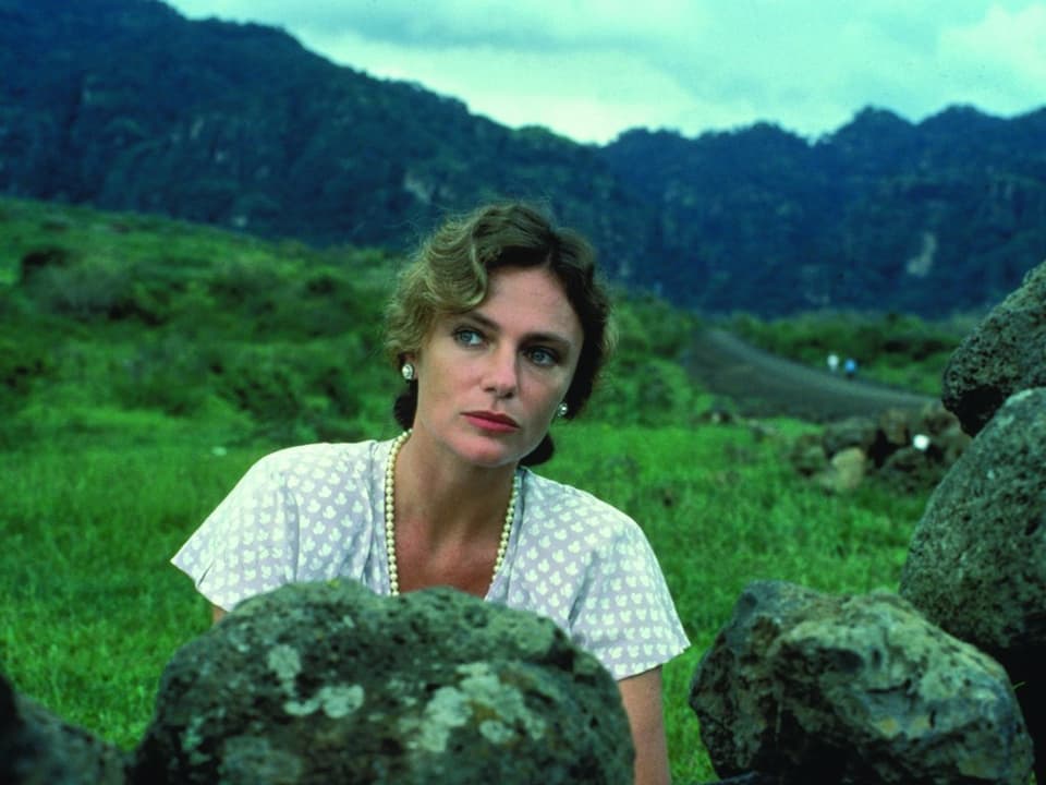 Eine geschminkte Frau in weisser Bluse blickt hinter einem grossen Stein hervor, dahinter eine Berglandschaft.