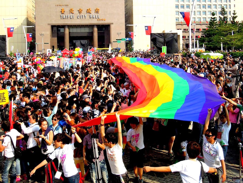 Viele Menschen an einer Parade, einige von ihnen halten ein mehrere Meter langes regenbogenfarbenes Tuch über den Köpfen.