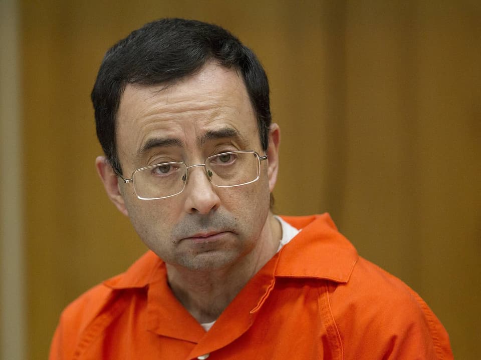 Mann in orangefarbener Häftlingskleidung mit Brille in einem Gerichtssaal.
