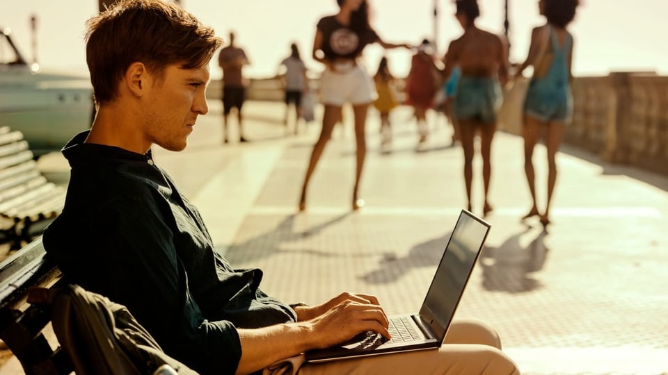 Junger, dunkelblonder Mann sitzt auf Bank, tippt am Laptop, hinter ihm eine sonnige Promenade.