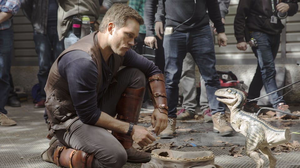 Schauspieler Chris Pratt am Filmset. Er kniet vor der einem kleinen, künstlichen Dinosaurier.