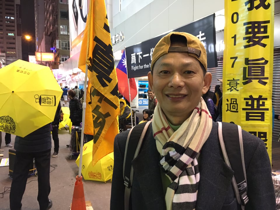 Ein junger Mann aus Hongkong steht vor gelben Fahnen und jemandem mit einem gelben Regenschirm.