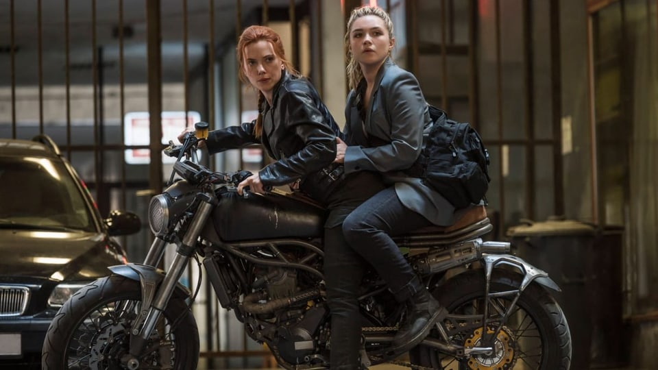 Zwei Frauen auf Motorrad, vorne eine Frau mit rotem, zusammengebundenem Haar, Lederjacke, hinten in Jeans blonde Frau.