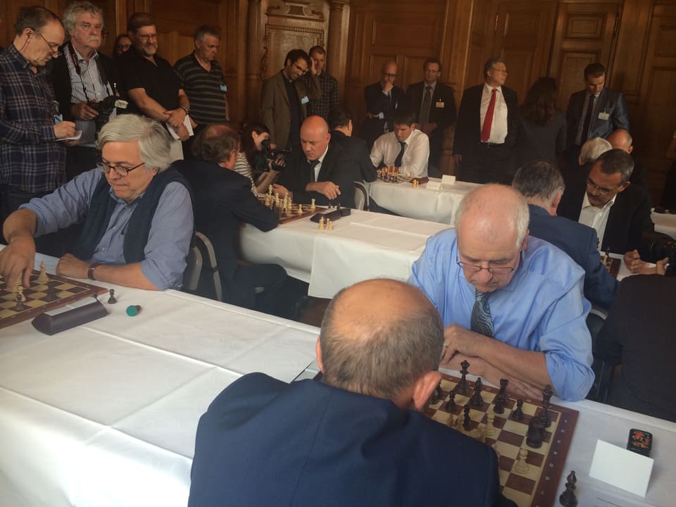Drei Tische, weisse Tischtücher, sechs Schachpartien mit zwölf Spielern sind im Gang. Rundherum Journalisten und Fotografen, zuhinterst steht Lombardi.