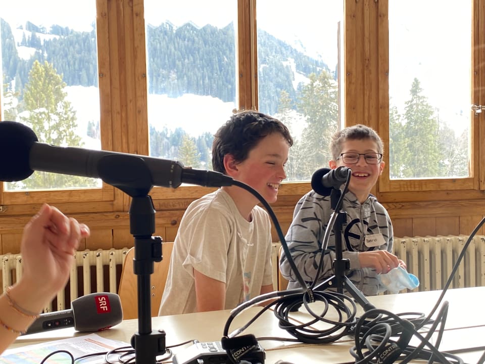 Zwei Jungen sitzen vor einem Mikrofon und lachen. Im Hintergrund sieht man durch das Fenster die Berge.