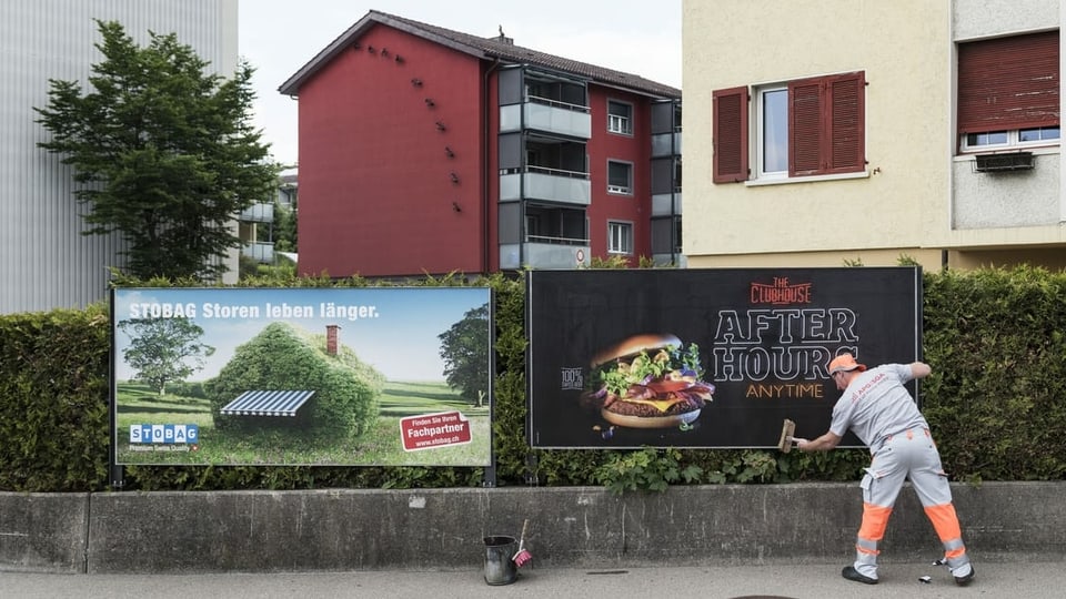Ein Mann klebt neue Plakate auf zwei Plakatwände in einer städtischen Umgebung.