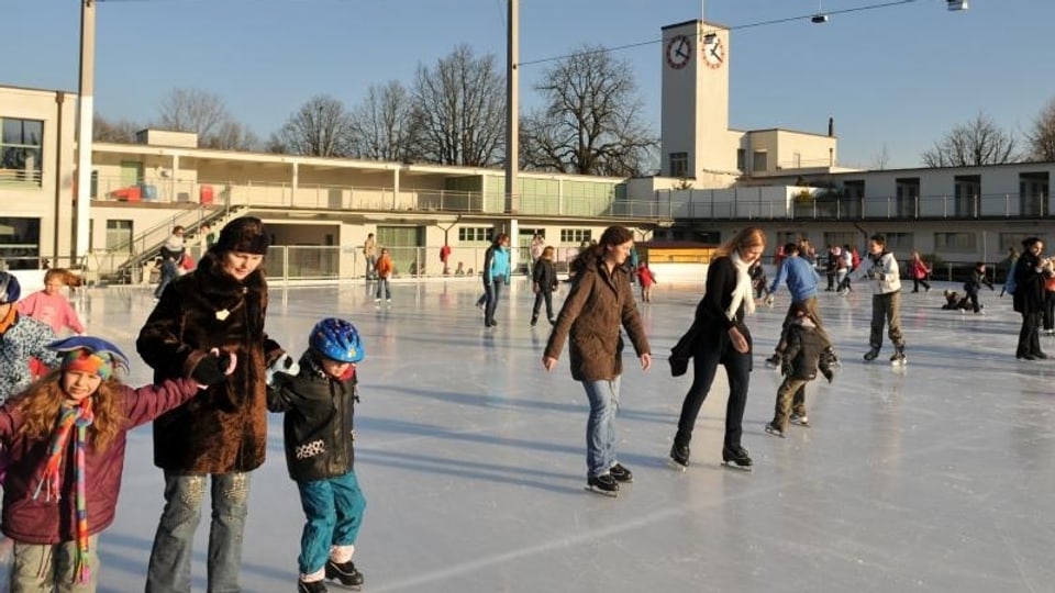 Eine Mutter führt Sohn und Tochter an den Händen übers Eis. Zahlreiche andere Schlittschuhläufer sind zu sehen. Im Hintergrund der Turm des Eglisee mit der Uhr.