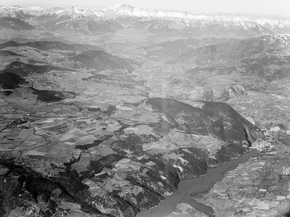 Ein See, Hügellandschaft und im Hintergrund Berge aus der Luft fotografiert.