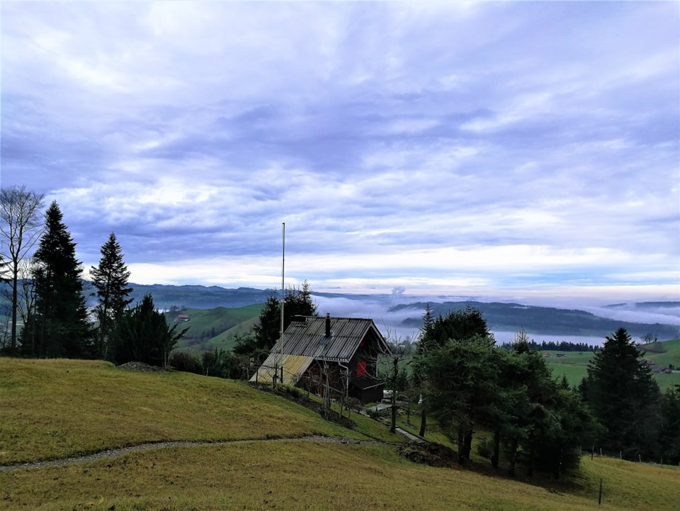 Hügelige Landschaft im Luzern, eine Hütte zwischen zwei Bäumen, im Hintergrund Nebelfeder, darüber  tauch die Berge aus dem Nebelgtrau. Der Himmel ist von vielen Wolken überzogen.