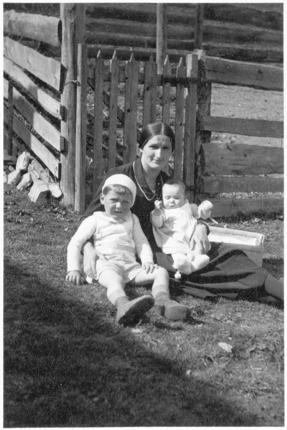 Schwarzweiss-Fotografie einer Frau mit zwei Kindern im Gras vor einem Zaun.