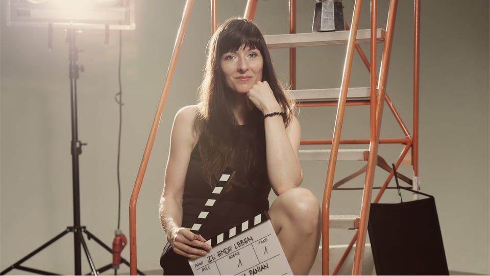 Frau in schwarzem Kleid sitzt mit Filmklappe in der Hand auf Leiter.