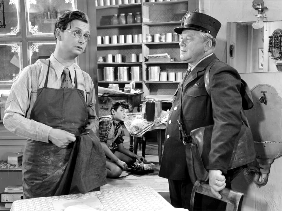 Ein uniformierter Mann mit Umhängetasche steht in einem Laden an der Verkaufstheke. Dahinter steht ein junger Mann in Schürze und scheint einen Gegenstand mit einem Tuch zu polieren. Im Hintergrund ist noch ein junger Mann zu erkennen.