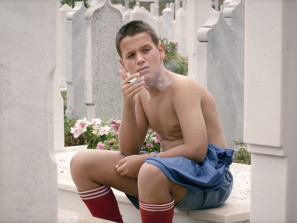 Eine Junge mit Fusball-Stulpen und nacktem Oberkörper sitzt rauichend auf einem Grabstein.