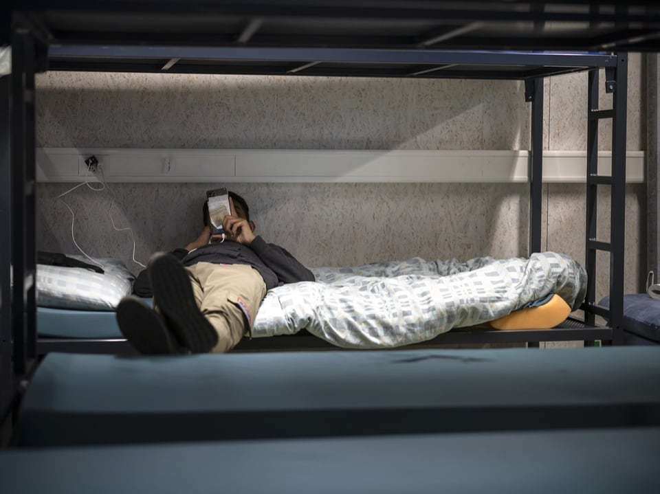 Ein Mann liegt mit einem Smartphone in der Hand auf einem Bett in einem Bundesayslzentrum.