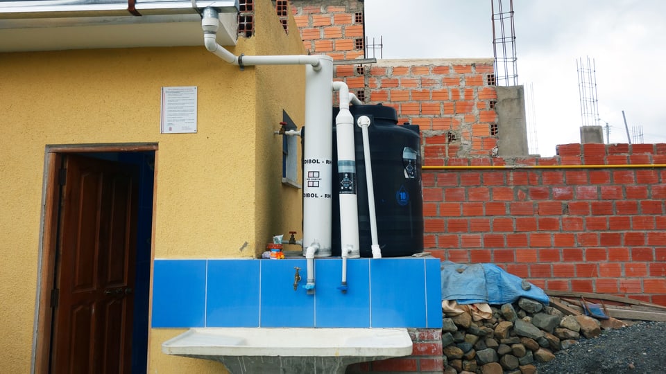 Eine Regenwasser_Auffang-Anlage mit mehreren weissen Rohren und einem Bottich.
