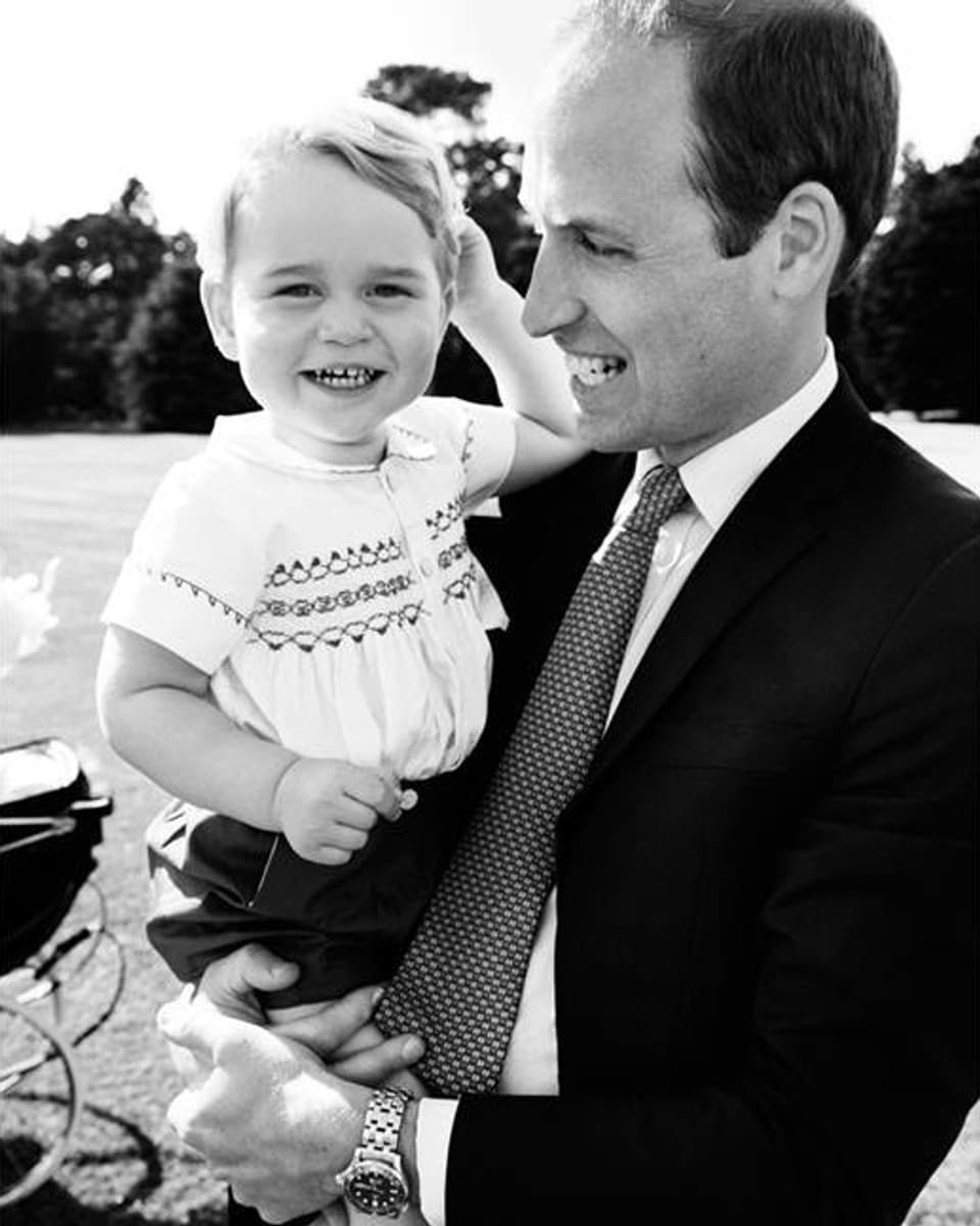 Prinz George lächelt in die Kamera, während ihn sein Vater William in den Armen hält.