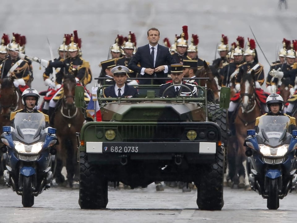 Macron steht auf dem Hintersitz eines fahrenden Militärfahrzeuges und wird von Sicherheitsleuten begleitet.