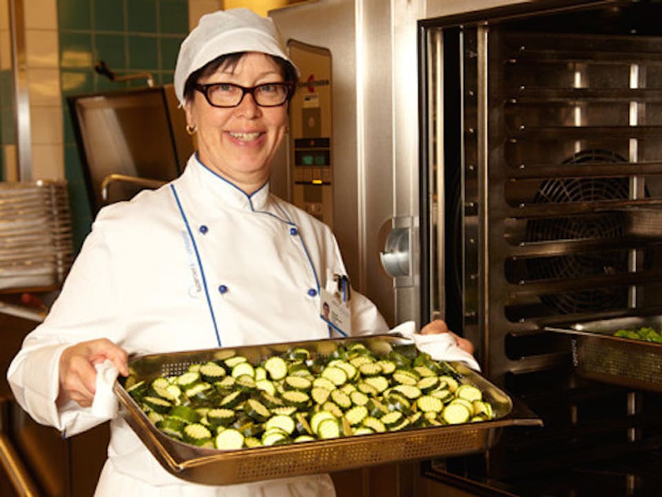 Frau in Kochbekleidung mit einem Ofenblech voll geschnittener Zucchini in der Hand