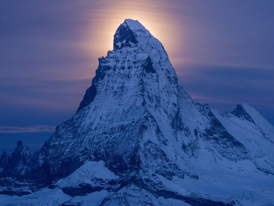 Verschneiter felsiger Berg, der oben erleuchtet wird. Der Himmel ist bedeckt. 