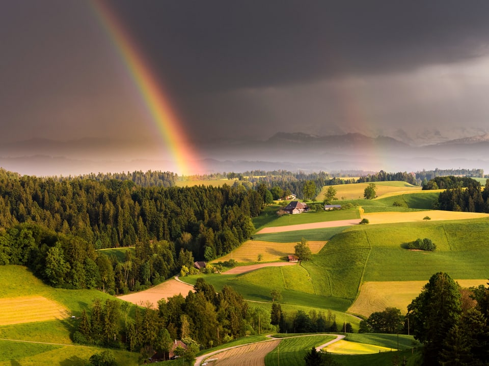 Regenbogen mit dunklen Wolken über einer grünen Hügellandschaft.