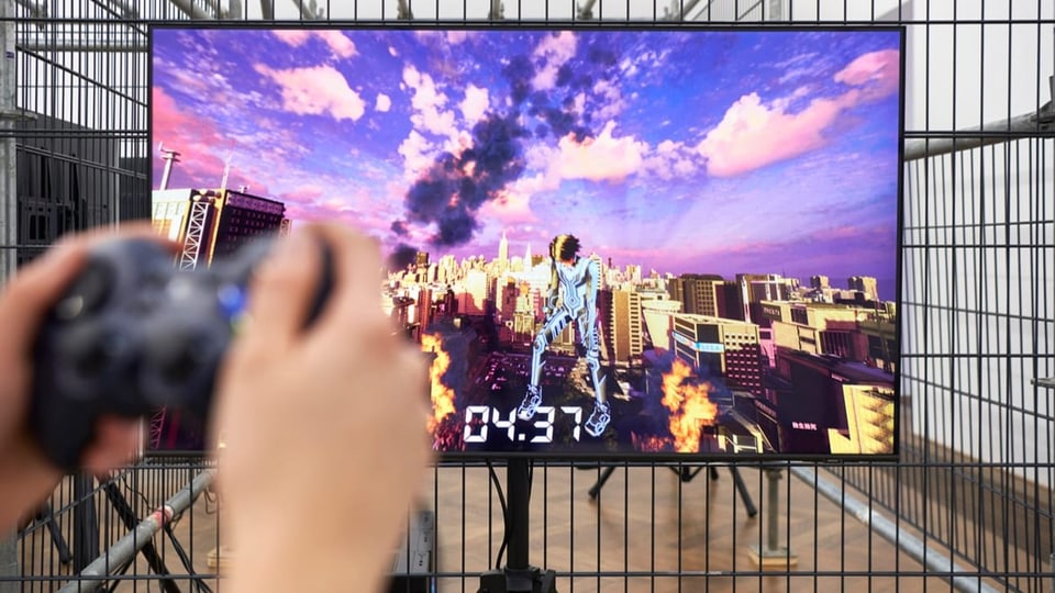 Links verschwommen eine Gamekonsole in den Händen, im Hintergrund scharf ein Bildschirm von einer Figur vor einer Stadt.